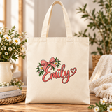 A natural cotton canvas tote bag with canvas handles, printed with a sample name in coral script with heart accents under a pink bow and white daisies, beside a jug of daisies and books.
