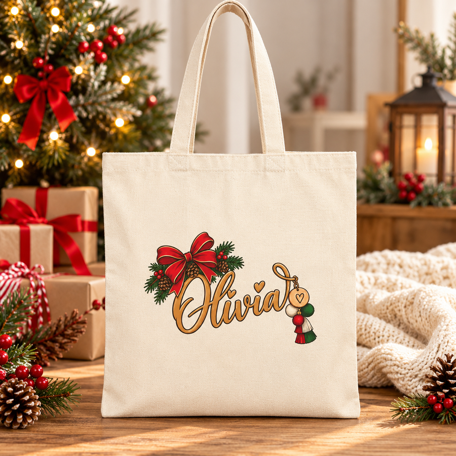A natural cotton canvas tote bag with canvas handles, printed with a sample name in gold script with a heart, under a red bow with pine and pinecones and a red, white and green tassel charm, in front of a Christmas tree and wrapped presents.