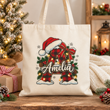 A natural cotton canvas tote bag with canvas handles, printed with a red and black buffalo plaid initial wearing a Santa hat with a red bow, holly, pinecones and string lights, and a sample name in white script across it, in front of a frosted Christmas tree and presents.