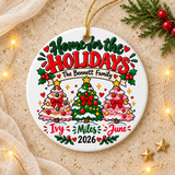A round white ceramic Christmas ornament on a gold string, printed with "Home for the Holidays" in green and red lettering above a banner with a sample family name, three decorated trees in pink and green, three sample first names and "2026".