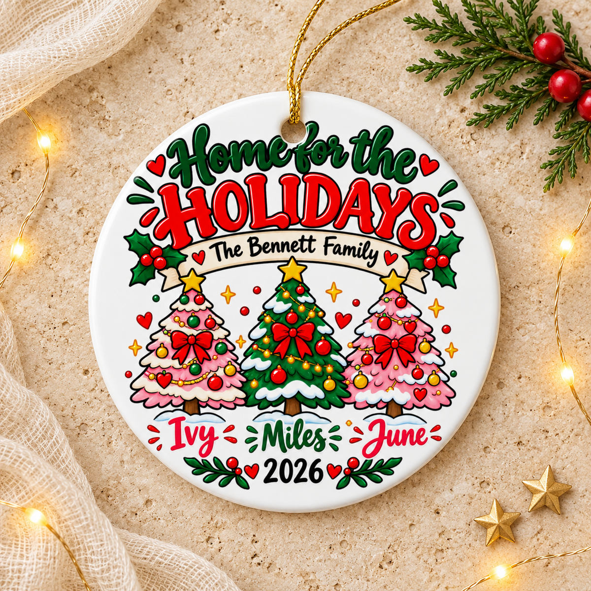 A round white ceramic Christmas ornament on a gold string, printed with "Home for the Holidays" in green and red lettering above a banner with a sample family name, three decorated trees in pink and green, three sample first names and "2026".