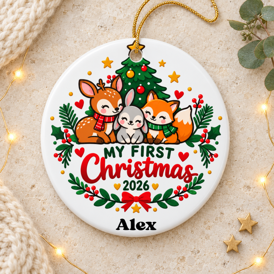 A round white ceramic Christmas ornament on a gold string, printed with a deer, a gray bunny and a fox in knit scarves cuddled under a decorated Christmas tree, above "My First Christmas" in green and red lettering, "2026", a holly garland with a red bow and a sample baby name.