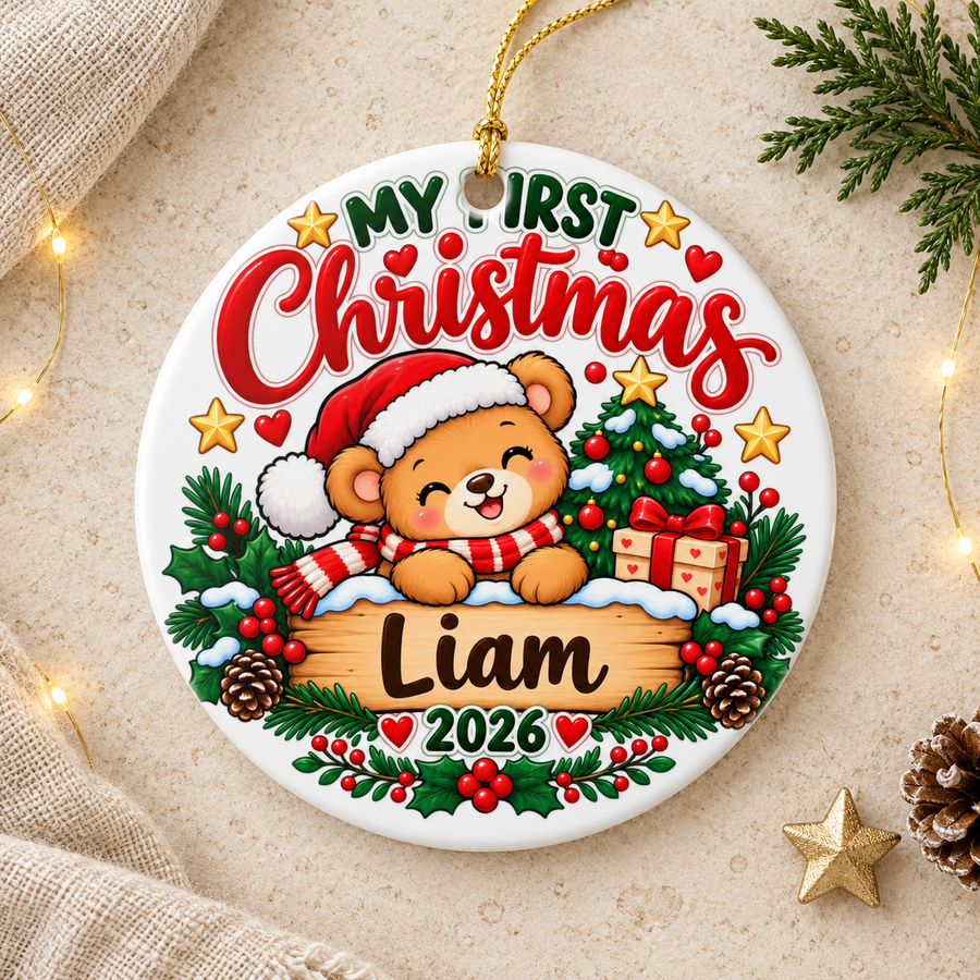 A round white ceramic Christmas ornament on a gold string, printed with "My First Christmas" in green and red lettering over a smiling teddy bear in a Santa hat and striped scarf, peeking over a wooden sign with a sample baby name and "2026", with a decorated tree, a present, pinecones and holly.