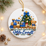 A round white ceramic Christmas ornament on a gold string, printed with a blue and cream toy train carrying presents and a teddy bear in a blue bow past a decorated tree, above "My First Christmas", a sample baby name in blue script and "2026".
