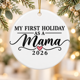 A round white ceramic Christmas ornament on a gold string, printed with "My First Holiday as a" in black lettering above "Mama" in large black script with flourishes, a small pink heart and "2026".