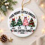 A round white ceramic Christmas ornament on a gold string, printed with "Our Merry Little Forest" in black lettering over four decorated trees, one pink, each above a sample first name, with a sample family name and "2026" below, plus holly, stars and hearts.