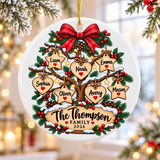 A round white ceramic Christmas ornament on a gold string, printed with a snowy tree under a red plaid bow, hung with wooden hearts and stars carrying sample first names, above a wooden sign with a sample family name and "2026", with berries, pinecones and lights.