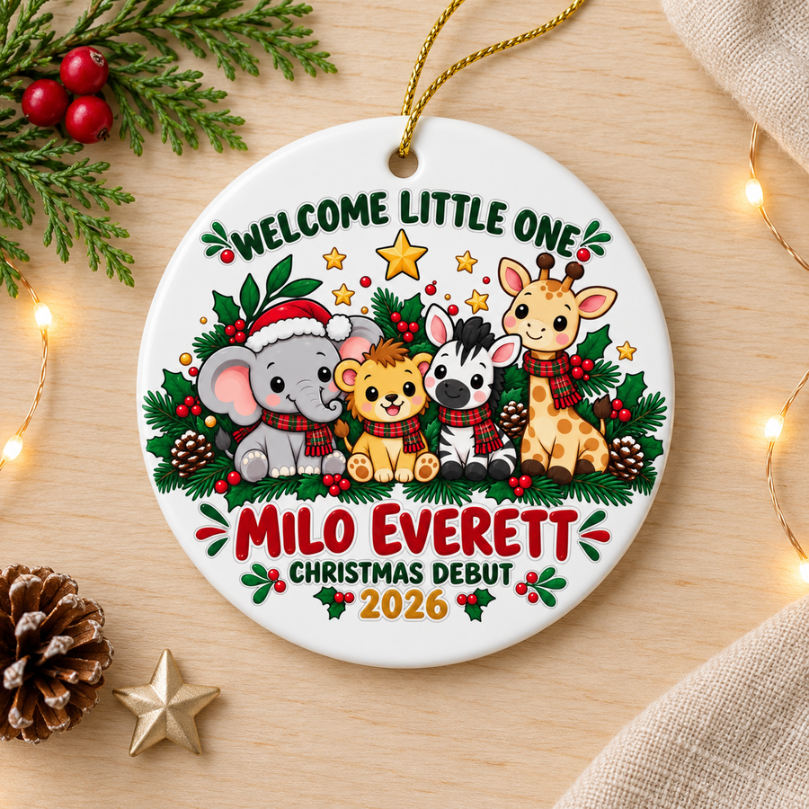 A round white ceramic Christmas ornament on a gold string, printed with "Welcome Little One" in green lettering over a baby elephant in a Santa hat, a lion cub, a zebra and a giraffe in plaid scarves among evergreen and pinecones, above a sample baby name in red, "Christmas Debut" and "2026".