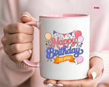 A white ceramic mug with a colored handle and interior, printed with "Happy Birthday" in pink and blue script above an orange "TO YOU" banner, with balloons, party bunting, party hats and confetti.