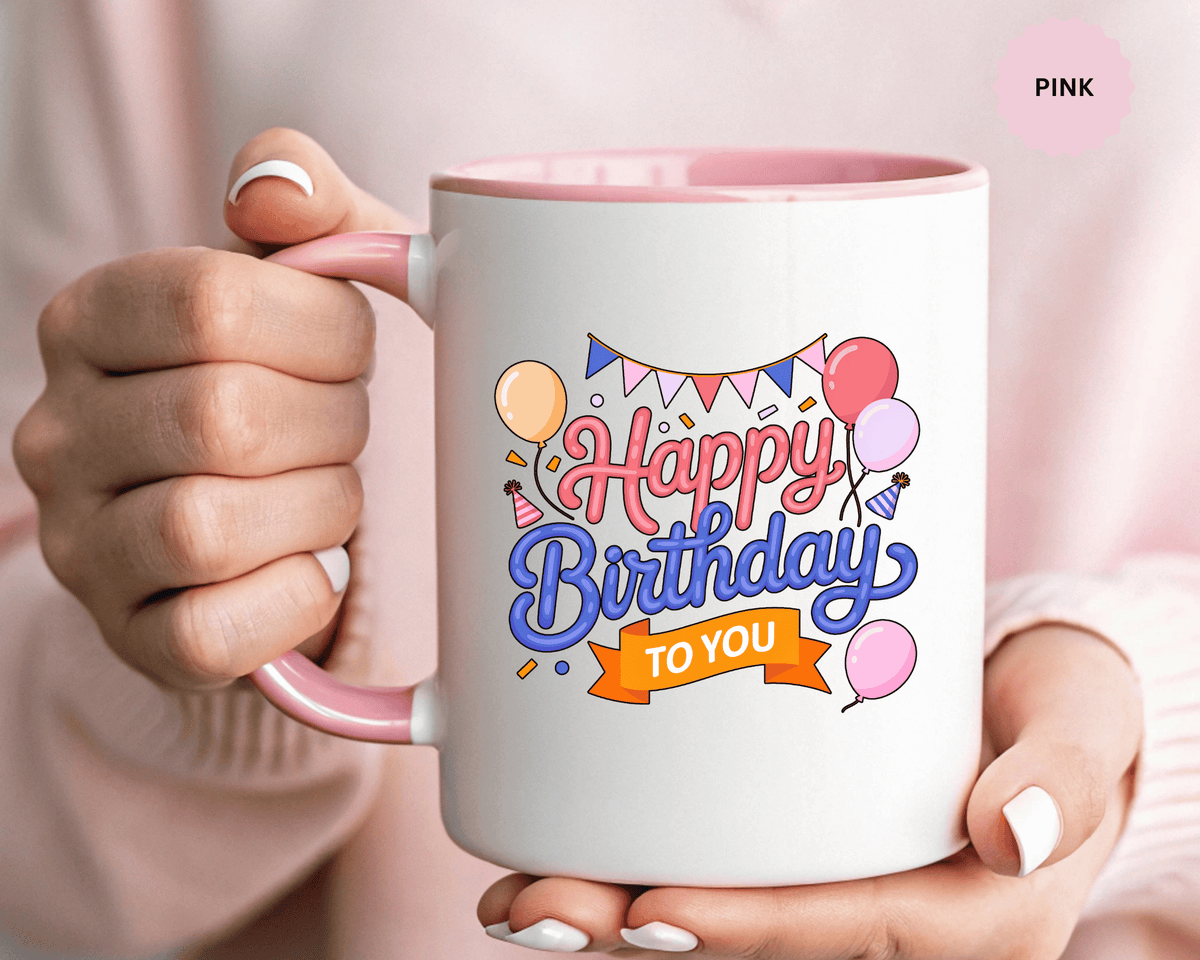 A white ceramic mug with a colored handle and interior, printed with "Happy Birthday" in pink and blue script above an orange "TO YOU" banner, with balloons, party bunting, party hats and confetti.