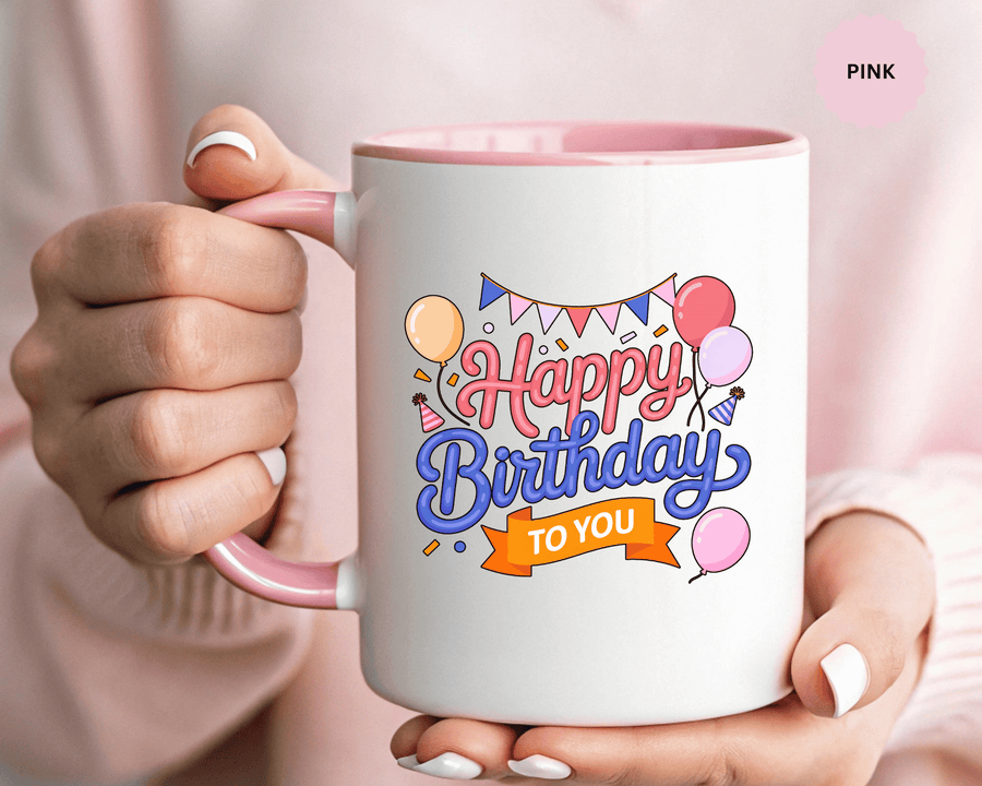 A white ceramic mug with a colored handle and interior, printed with "Happy Birthday" in pink and blue script above an orange "TO YOU" banner, with balloons, party bunting, party hats and confetti.