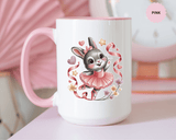 A white ceramic mug with a colored handle and interior, printed with a gray bunny with a pink bow in a pink tutu and ballet shoes, dancing among pink ribbons, hearts, stars and small flowers.