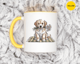 A white ceramic mug with a colored handle and interior, printed with a fluffy cream and tan puppy holding white daisies in its mouth, sitting on a green gingham blanket.