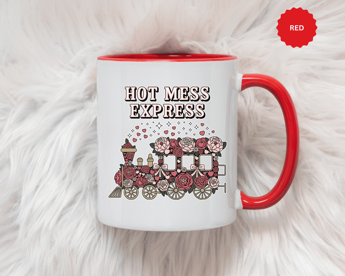 A white ceramic mug with a colored handle and interior, printed with "HOT MESS EXPRESS" in cream and black lettering above a train covered in pink and red roses with bows, hearts and sparkles.