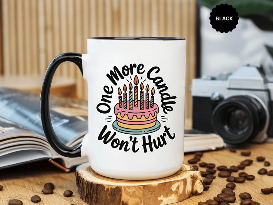 A white ceramic mug with a colored handle and interior, printed with "One More Candle Won't Hurt" in black script around a pink and yellow layered birthday cake with striped lit candles.