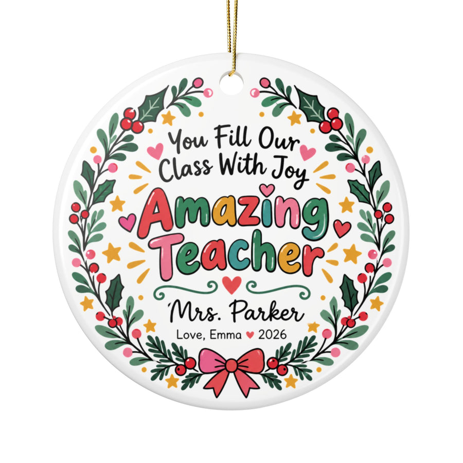 A round white ceramic Christmas ornament on a gold string, printed with "You Fill Our Class With Joy" and "Amazing Teacher" in colorful lettering inside a holly and berry wreath with a pink bow, with a sample teacher name and a "Love" signature with "2026".