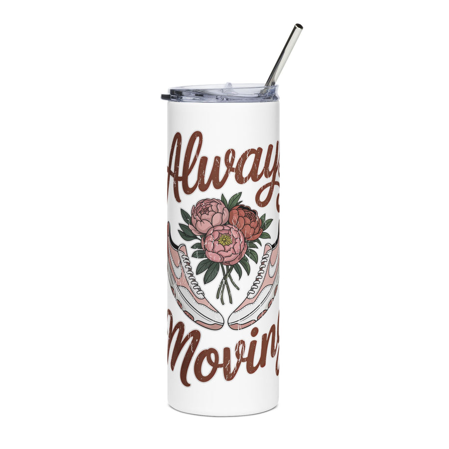 A white insulated tumbler with a clear lid and metal straw sits on a wooden table in a bright, pink-themed room. The tumbler is printed with brown script text reading 'Always Moving' and an illustration of pink flowers and white sneakers.