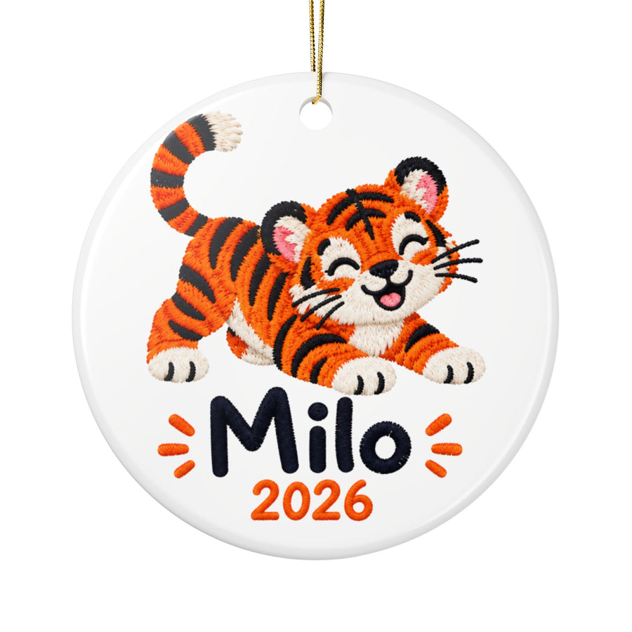 A round white ceramic Christmas ornament on a gold string, printed with a smiling orange and black striped tiger cub in an embroidery style, above a sample child's name in black and "2026" in orange.