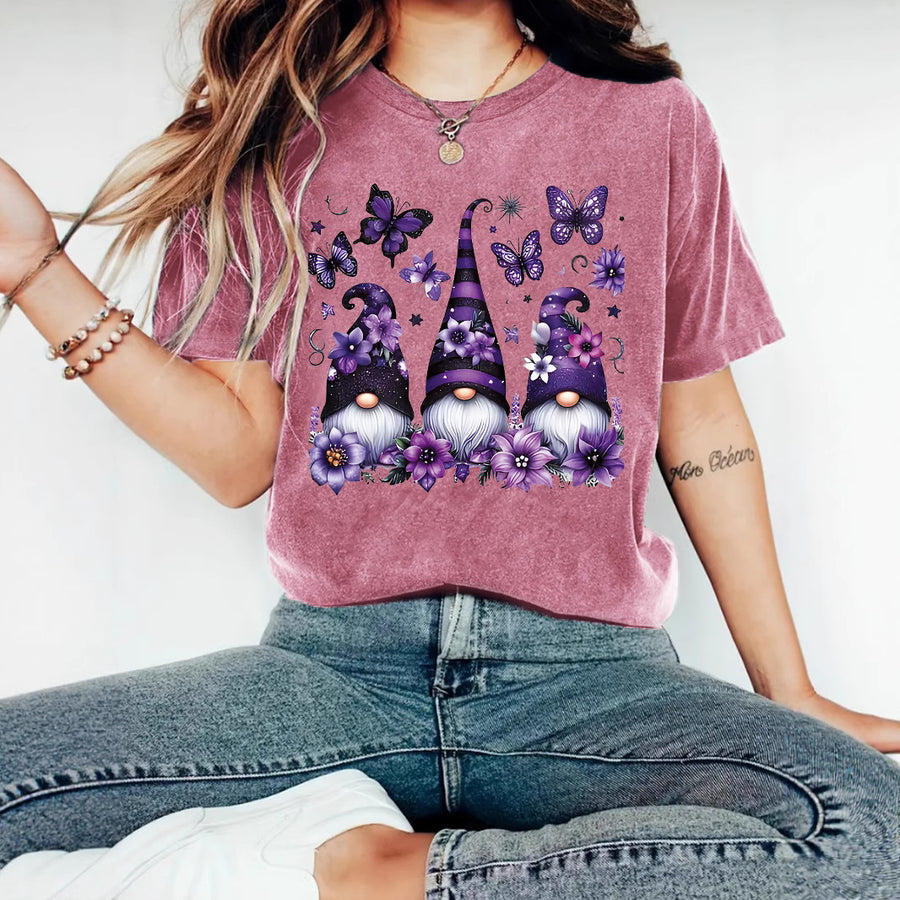 A plum T-shirt printed with three gnomes in purple patterned hats among purple flowers and butterflies.