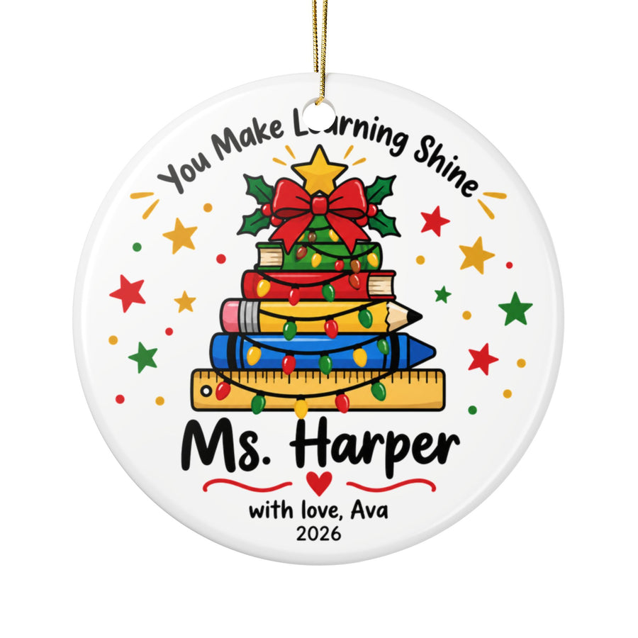 A round white ceramic Christmas ornament on a gold string, printed with "You Make Learning Shine" above a Christmas tree made of stacked books, a pencil and a ruler wrapped in lights, with a sample teacher name, "with love" signature and "2026".