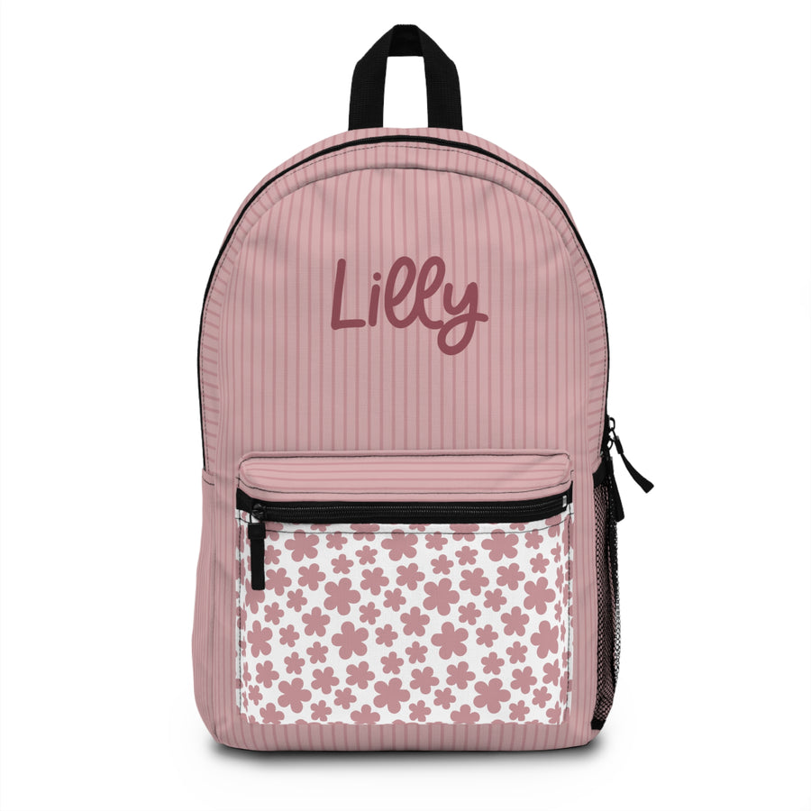 A pink backpack with thin pinstripes, a pink and white floral front pocket, black straps and zippers, and the sample name "Lilly" printed in dark pink script, in a bedroom.