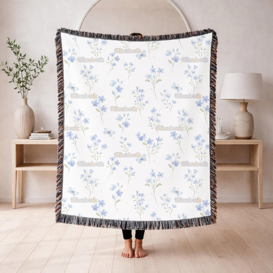 White fringed woven blanket held up in a living room, printed with delicate blue wildflowers and small butterflies, with the name Elizabeth repeated in cream retro lettering.