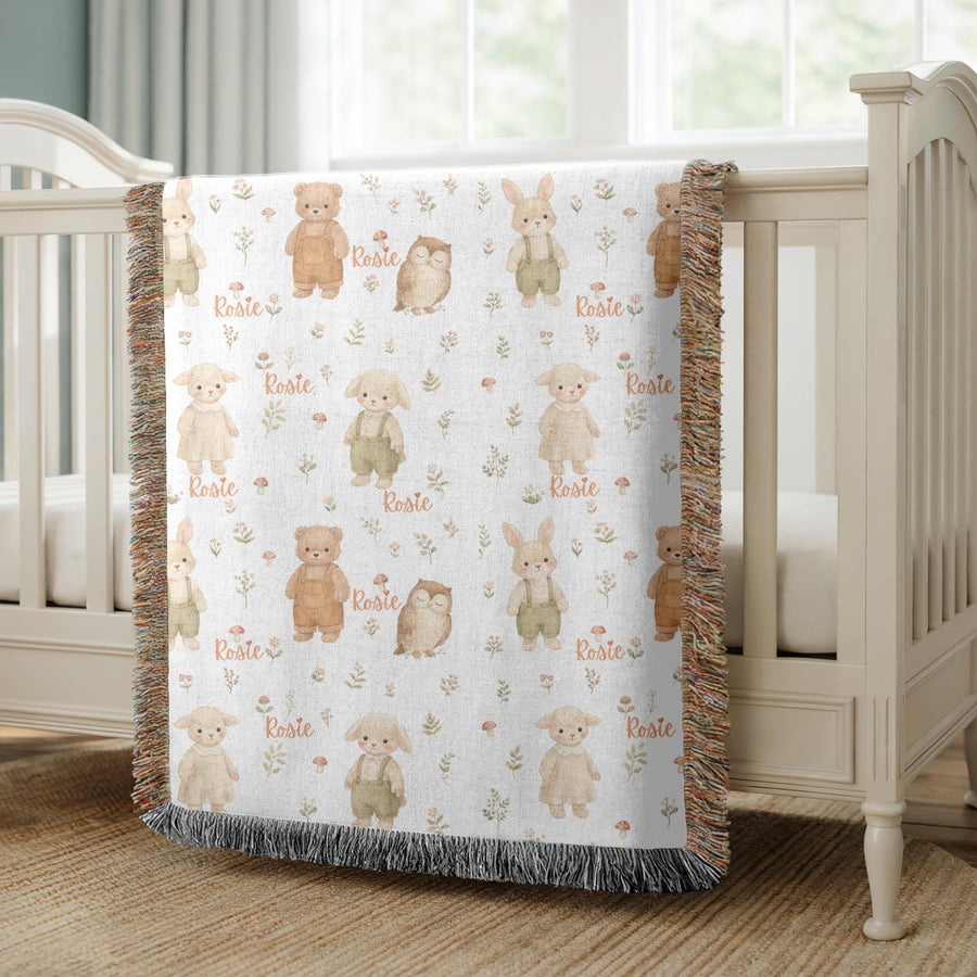 Fringed woven baby blanket printed with watercolour bunnies, bears in overalls, sleeping owls and lambs among red mushrooms and green sprigs, with the name Rosie in orange script.