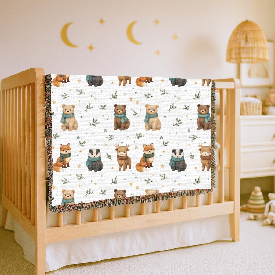 White fringed woven blanket printed with watercolour foxes, badgers, bears and a deer in pale teal scarves, set in rows among small gold stars and sprigs of green foliage.
