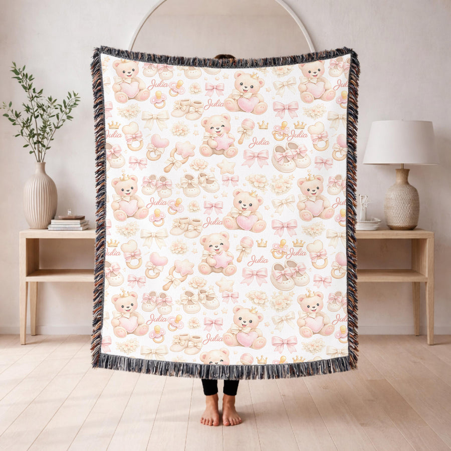 White fringed woven blanket over a cream crib, printed with pink crowned teddy bears holding hearts among bows, booties, rattles and pacifiers, with the name Julia in pink script.