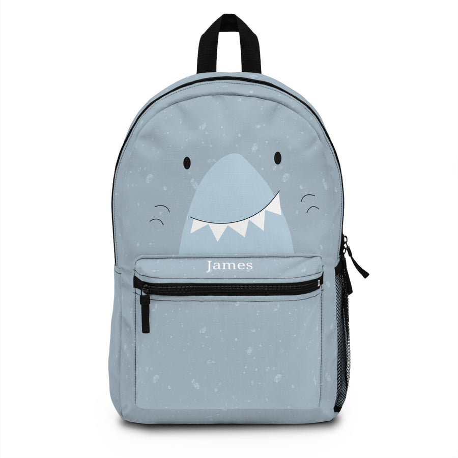 A kids backpack with a black top handle, front zip pocket and mesh side pocket, speckled light blue with a big grinning shark face and the sample name "James" in white on the pocket.
