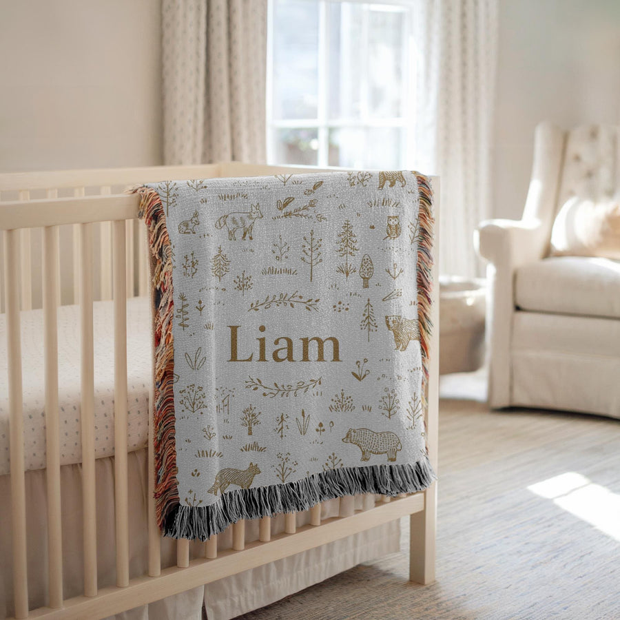White fringed woven baby blanket over a cream crib, covered in gold line drawn bears, foxes, owls and pine trees, with the name Liam in gold serif lettering across the centre.