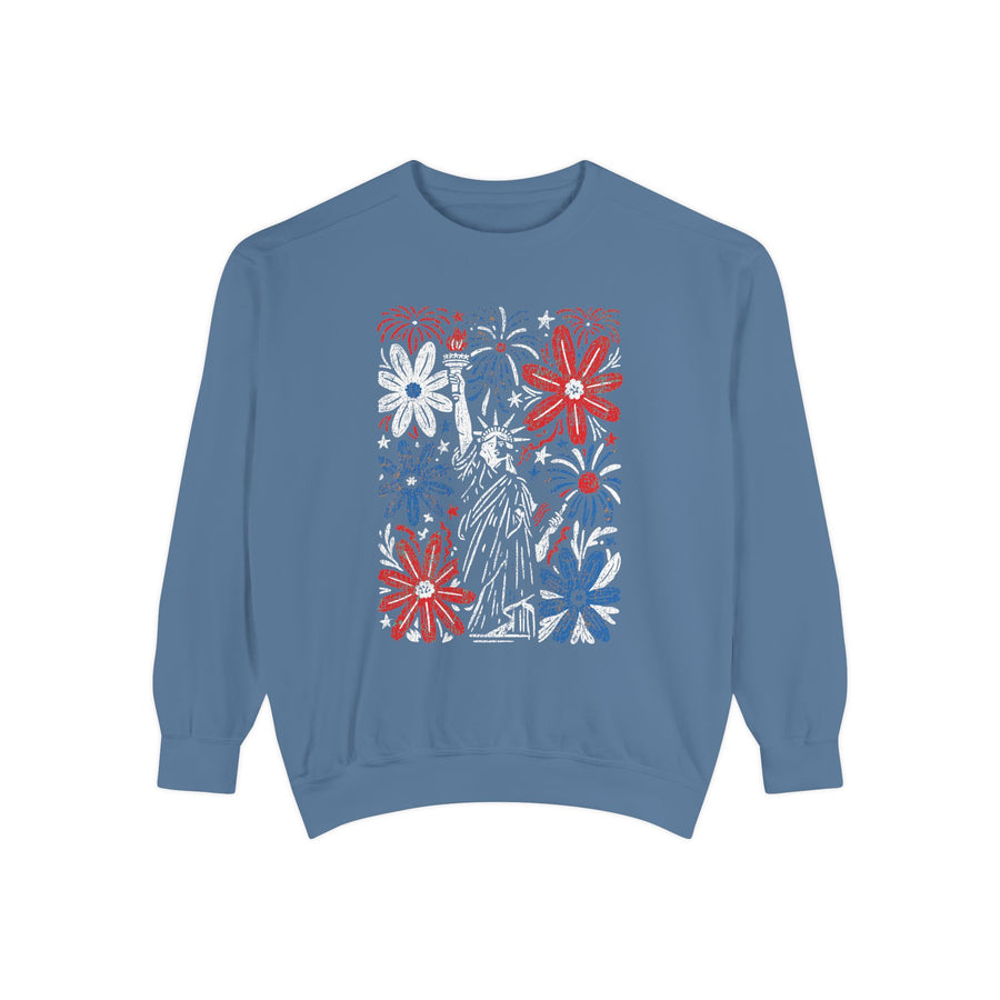 A blue crewneck sweatshirt printed with a panel of red, white and blue fireworks and flowers around the Statue of Liberty.