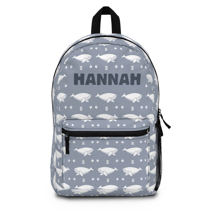 A kids backpack with a black top handle, front zip pocket and mesh side pocket, blue gray with an all-over pattern of white whales and small arrows and the sample name "HANNAH" in bold dark lettering.
