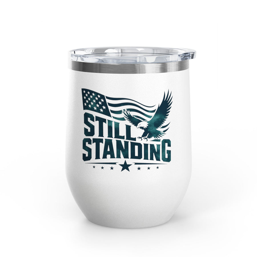 A white stainless steel stemless wine tumbler with a clear push-on lid, printed with "STILL STANDING" in dark green lettering with a soaring bald eagle, a waving American flag and stars.