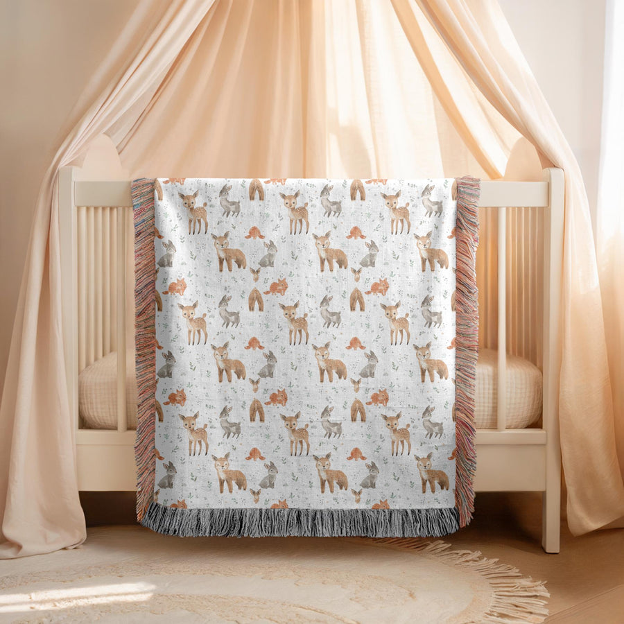 Fringed woven baby blanket held up in a living room, patterned all over with watercolour spotted fawns, foxes and grey rabbits among small orange mushrooms and pale green leaves.