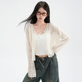 A woman wearing a cream open-knit cardigan patterned with fine pointelle rows and tied at the neck, worn over a white camisole with dark green wide leg jeans, her hair caught in the wind.