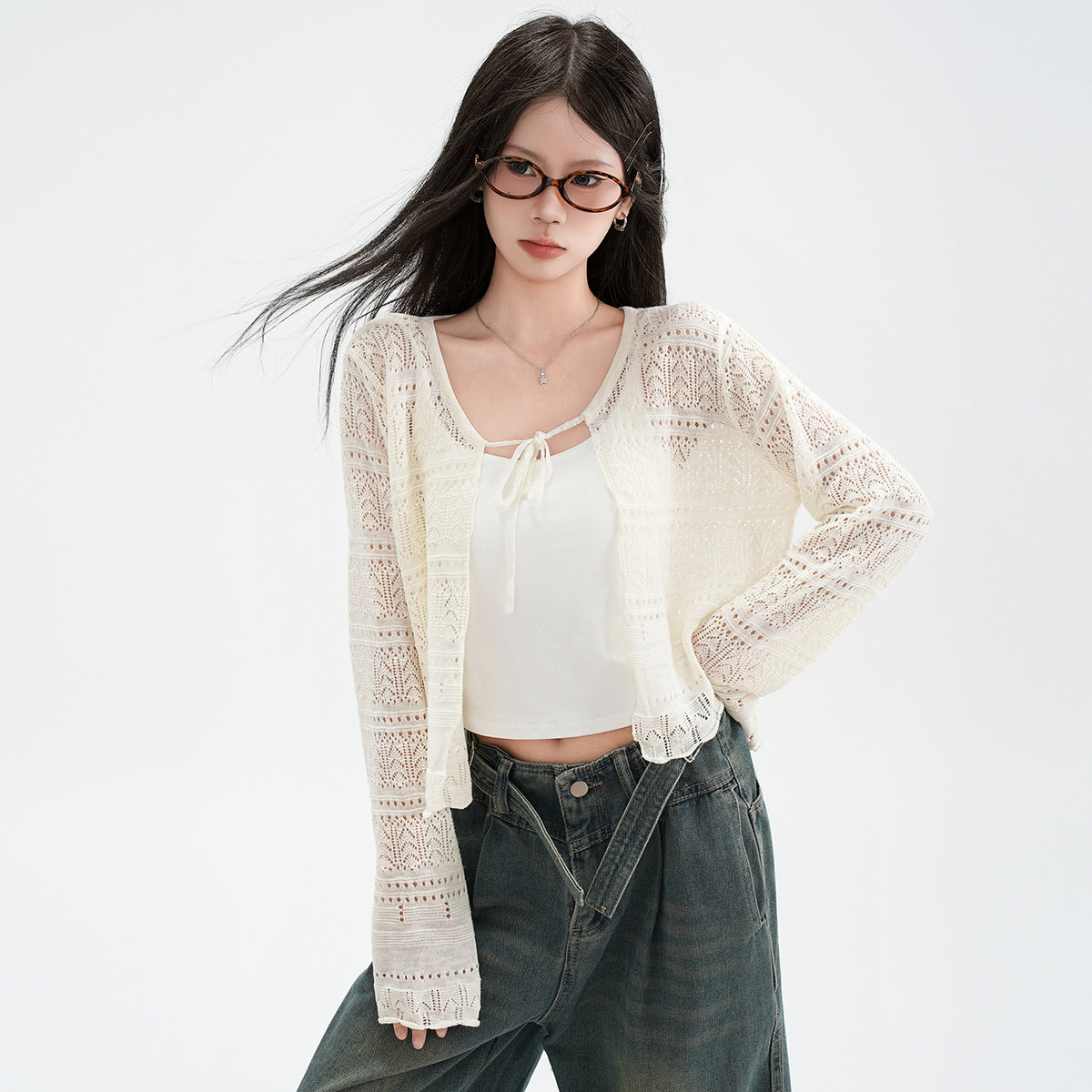 A woman wearing a cream open-knit cardigan patterned with fine pointelle rows and tied at the neck, worn over a white camisole with dark green wide leg jeans, her hair caught in the wind.