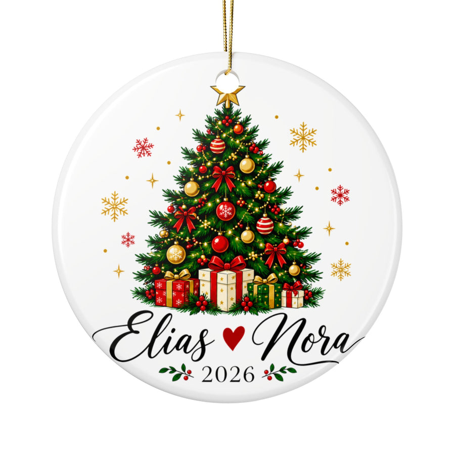 A round white ceramic Christmas ornament on a gold string, printed with a decorated Christmas tree with red and gold baubles, bows and wrapped presents under a gold star, above two sample names in black script joined by a red heart and "2026".