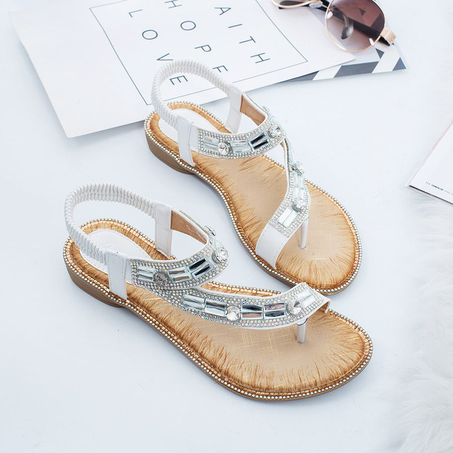 A pair of white flat sandals photographed from above, the T strap set with rows of clear and mirrored rhinestones and the heel held by an elasticated sling back, set on a white fur rug beside a card.