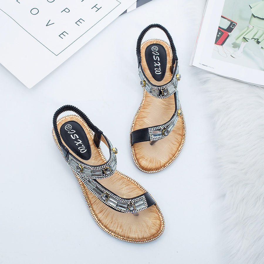 A pair of white flat sandals photographed from above, the T strap set with rows of clear and mirrored rhinestones and the heel held by an elasticated sling back, set on a white fur rug beside a card.