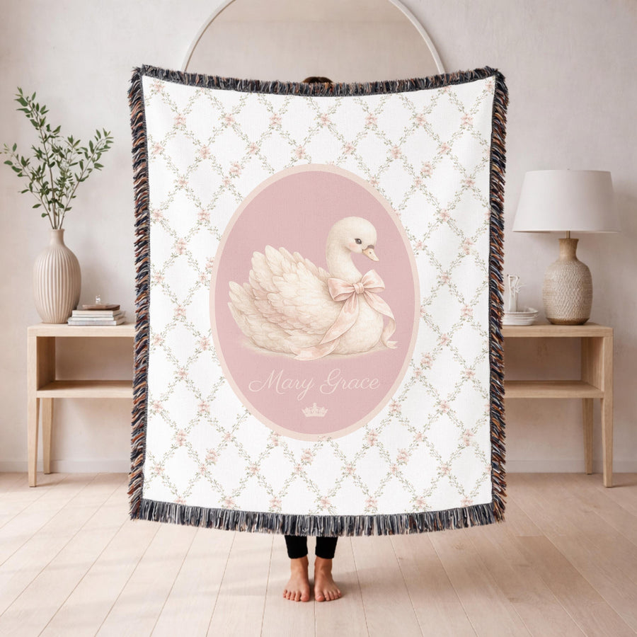 White fringed woven blanket on a cream crib, printed with a floral trellis and a central pink oval holding a white swan in a pale bow, with the name Mary Grace in cream script.