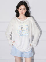 A woman wearing a loose cream long sleeved top printed at the chest with a pale blue line-drawn elephant above the hand-lettered word eleven, with pale blue shorts, both hands pushed into pockets.