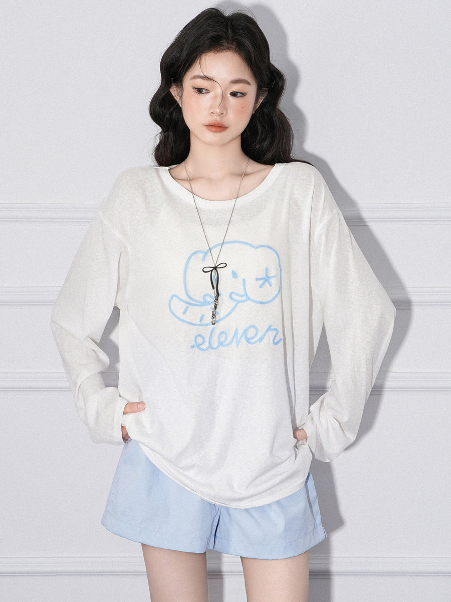 A woman wearing a loose cream long sleeved top printed at the chest with a pale blue line-drawn elephant above the hand-lettered word eleven, with pale blue shorts, both hands pushed into pockets.