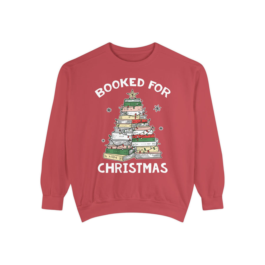 A woman wearing a light chambray blue sweatshirt with a graphic of a Christmas tree made of stacked books and the words 'BOOKED FOR CHRISTMAS'. She is holding a drink and sitting on a cozy couch.
