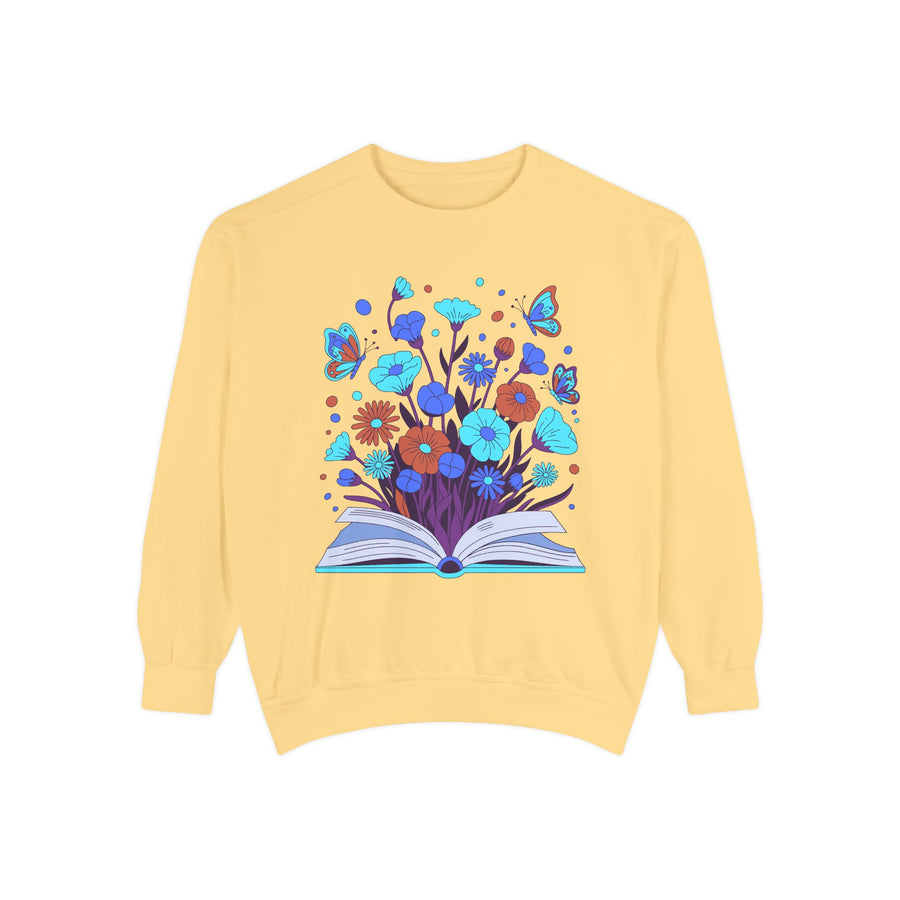 White sweatshirt printed with an open book from which blue and orange wildflowers spring upward, four butterflies rising through the blooms and a teal glow running along the open pages.