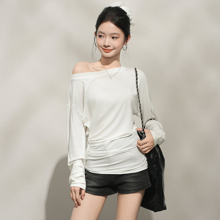 A woman wearing a cream long sleeved top with a wide boat neckline slipping off one shoulder, the soft fabric gathering at the waist and the sleeves cut full to the cuff.