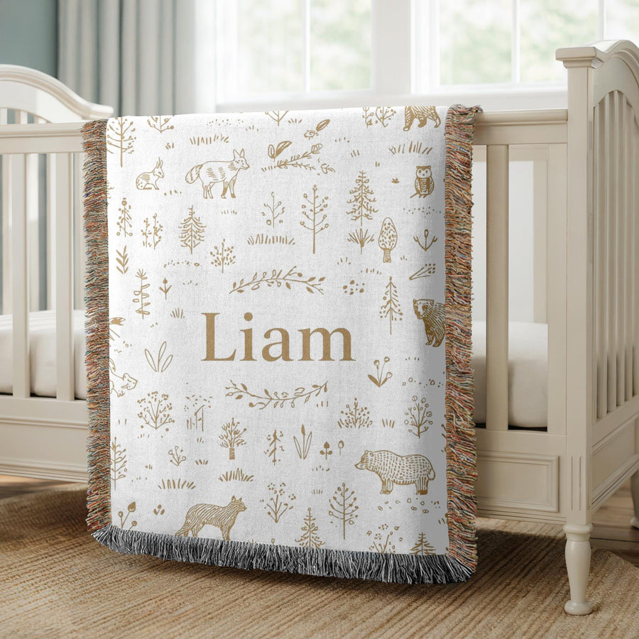 White fringed woven baby blanket over a cream crib, covered in gold line drawn bears, foxes, owls and pine trees, with the name Liam in gold serif lettering across the centre.