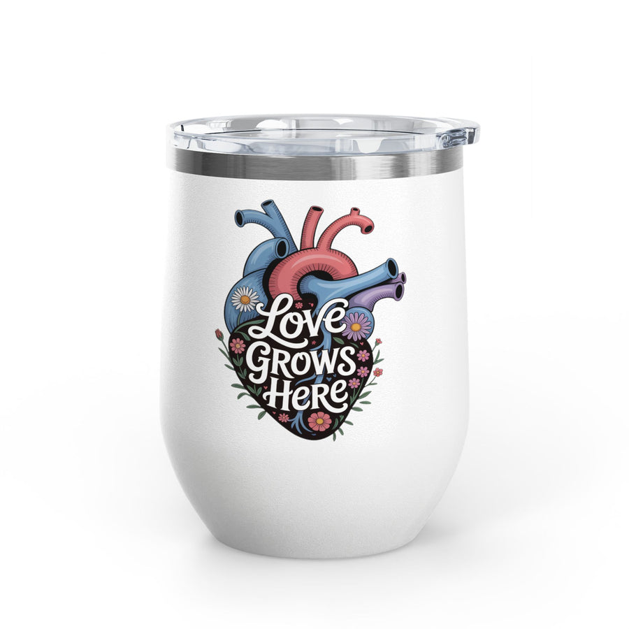 A white stainless steel stemless wine tumbler with a clear push-on lid, printed with "Love Grows Here" in white script on an anatomical heart in blue, red and purple, covered in daisies and small flowers.