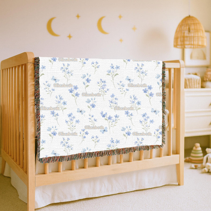 White fringed woven blanket held up in a living room, printed with delicate blue wildflowers and small butterflies, with the name Elizabeth repeated in cream retro lettering.