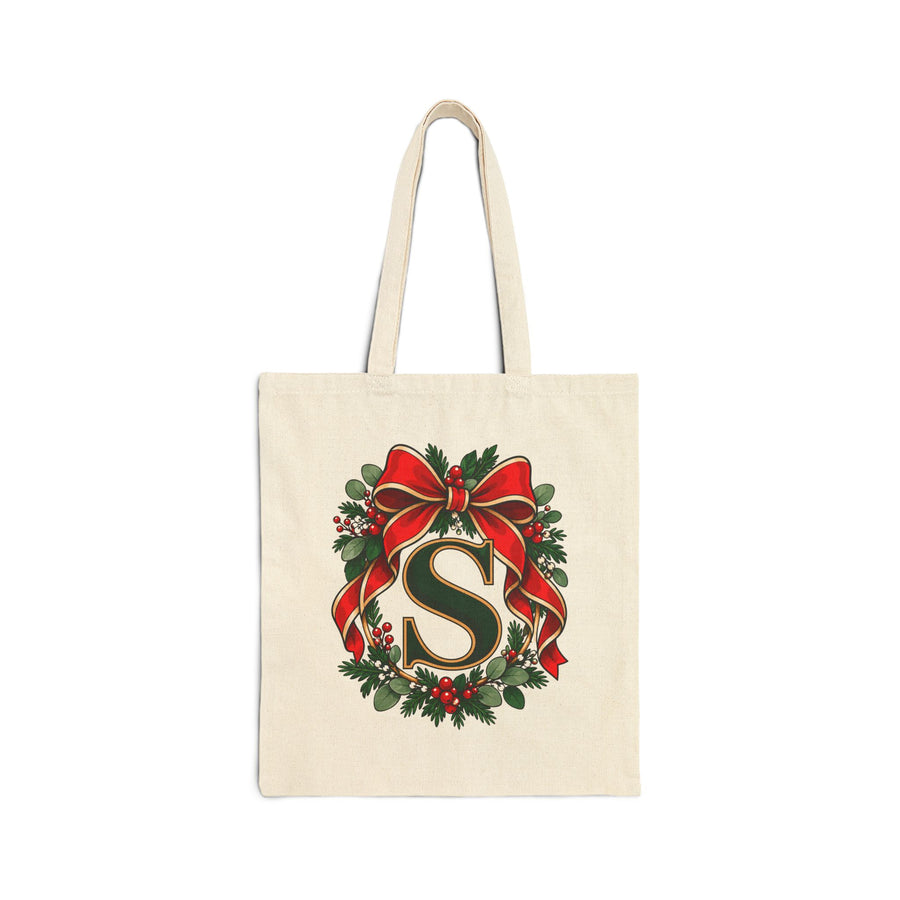 A natural cotton canvas tote bag with canvas handles, printed with a sample initial in dark green with a gold outline inside an evergreen wreath with red berries and a large red ribbon bow, in front of a frosted Christmas tree and presents.